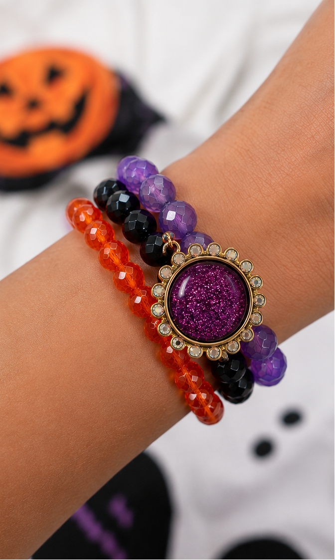Three Halloween-themed beaded stretch bracelets displayed on a white surface, with one shown worn on a person's wrist. The bracelets feature purple, black, and orange faceted beads with a central glittery purple oval charm surrounded by gold-tone detailing and small clear rhinestones.