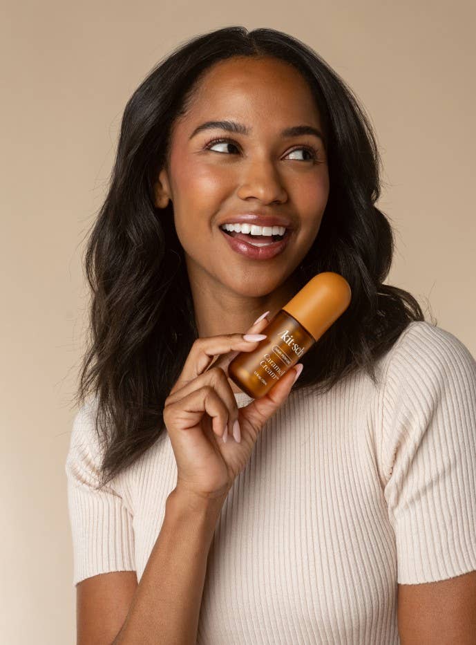 A smiling woman with long dark hair holds a small amber bottle against a warm beige background. The bottle appears to be a skincare or cosmetic product, and the brand name is partially visible on the label.