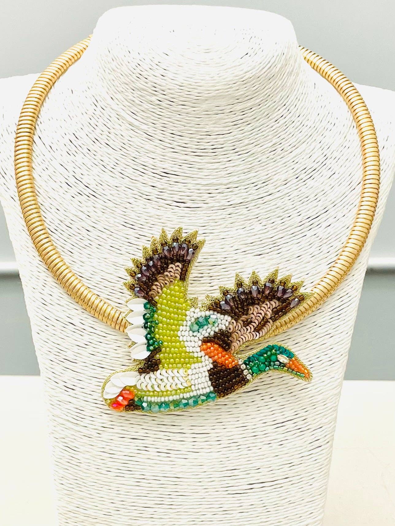 A gold-tone beaded necklace displayed on a white textured mannequin bust, featuring a large duck-shaped pendant with green, brown, white, and orange beadwork.