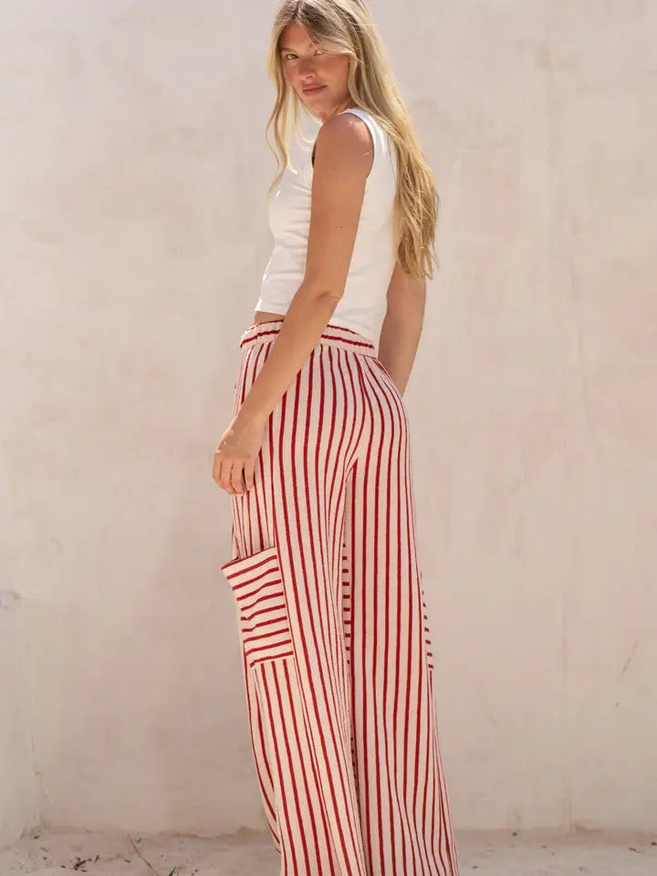A woman wearing red and white vertically striped wide-leg pants with a matching striped pocket, paired with a white sleeveless top, standing against a textured beige wall.