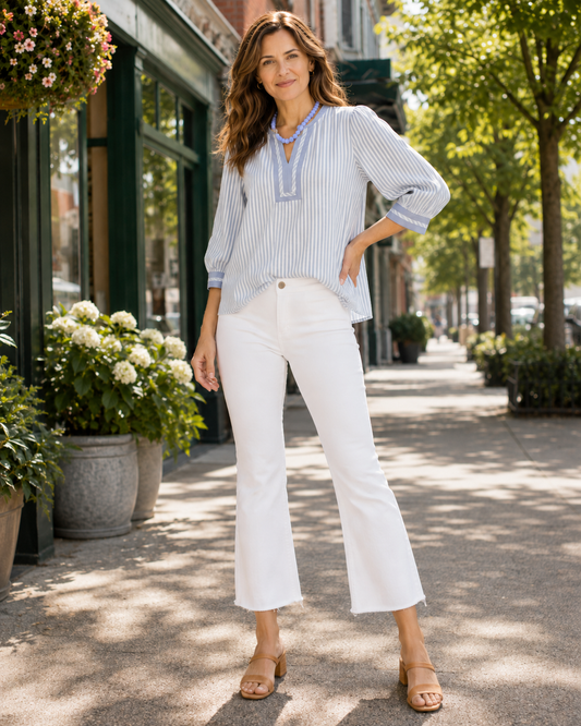 A woman stands on a sunlit city sidewalk wearing a light blue and white vertically striped long-sleeve blouse with a split neckline, paired with white cropped pants and tan heeled sandals.