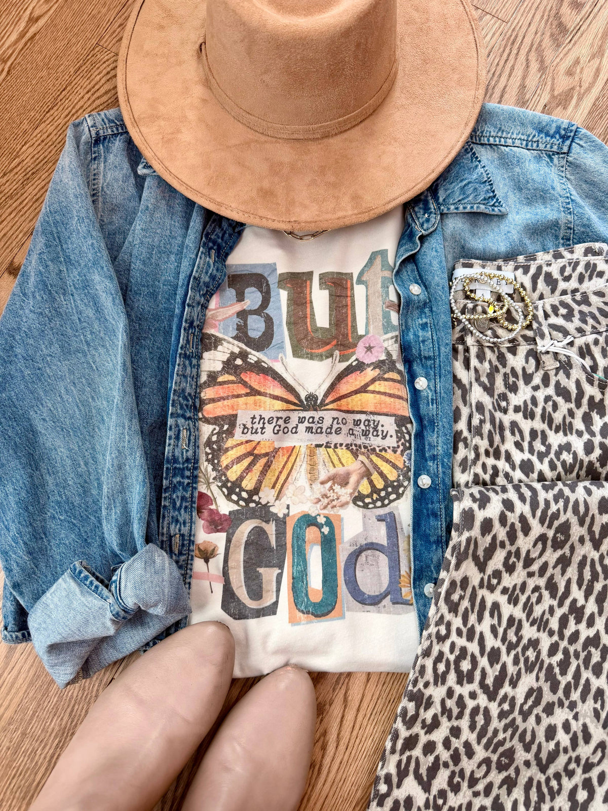 Person wearing a denim jacket, white graphic t-shirt with text and butterfly design, and brown hat on a wooden surface.