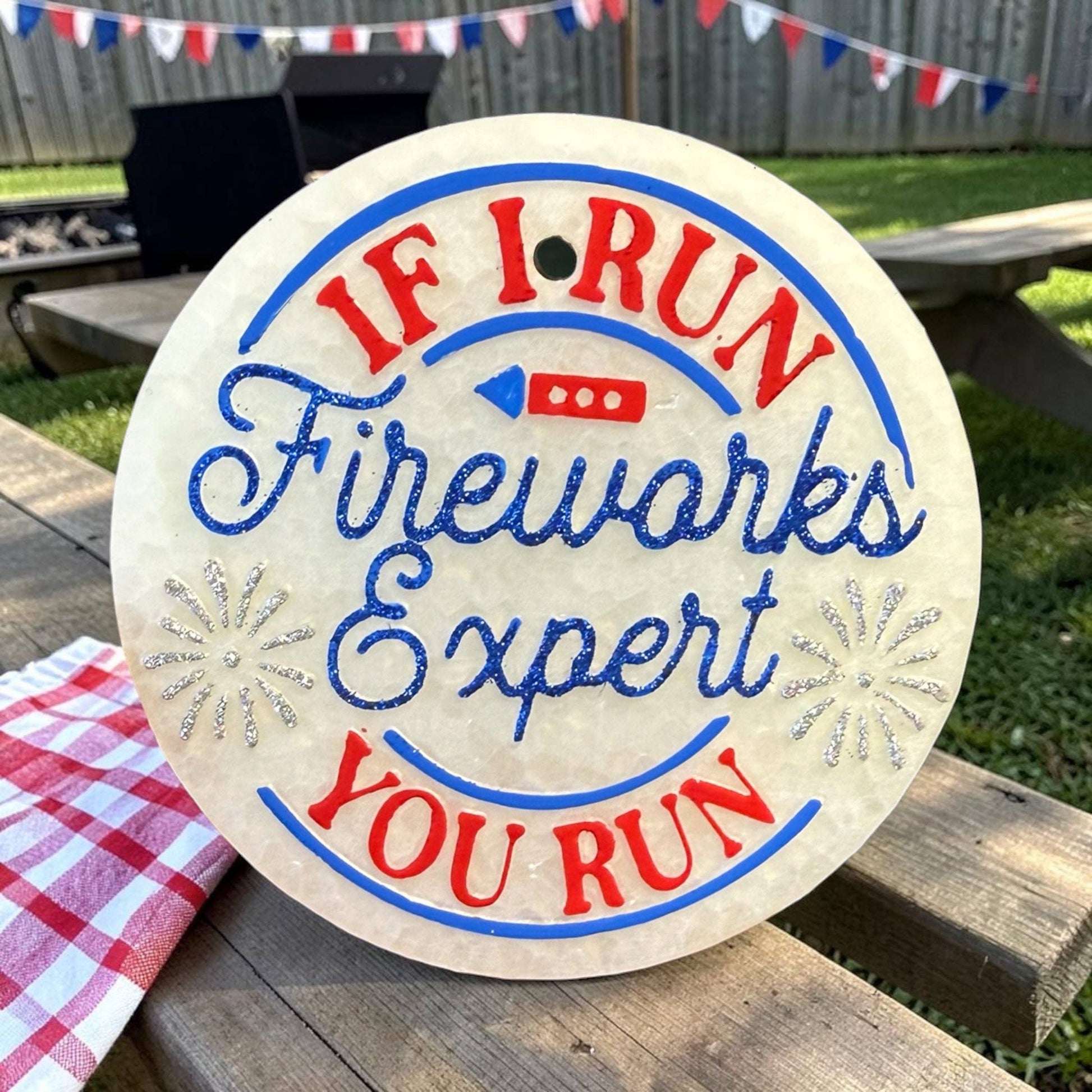 Round sign with text 'If I Run Fireworks Expert, You Run' on a wooden surface outdoors.