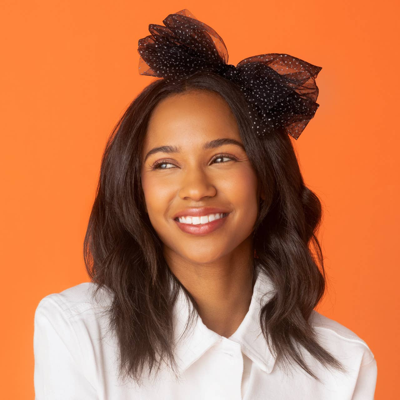 A smiling woman with dark wavy hair wearing a black sheer tulle bow headband with small white polka dots, set against a solid bright orange background.