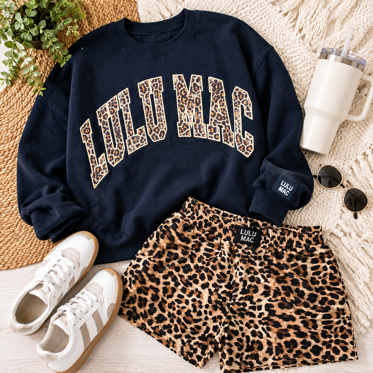 A styled flat-lay outfit featuring a navy blue sweatshirt with leopard-print 'LULU MAC' lettering, matching leopard-print shorts, white sneakers, sunglasses, and a white tumbler on a woven mat and knit blanket background.
