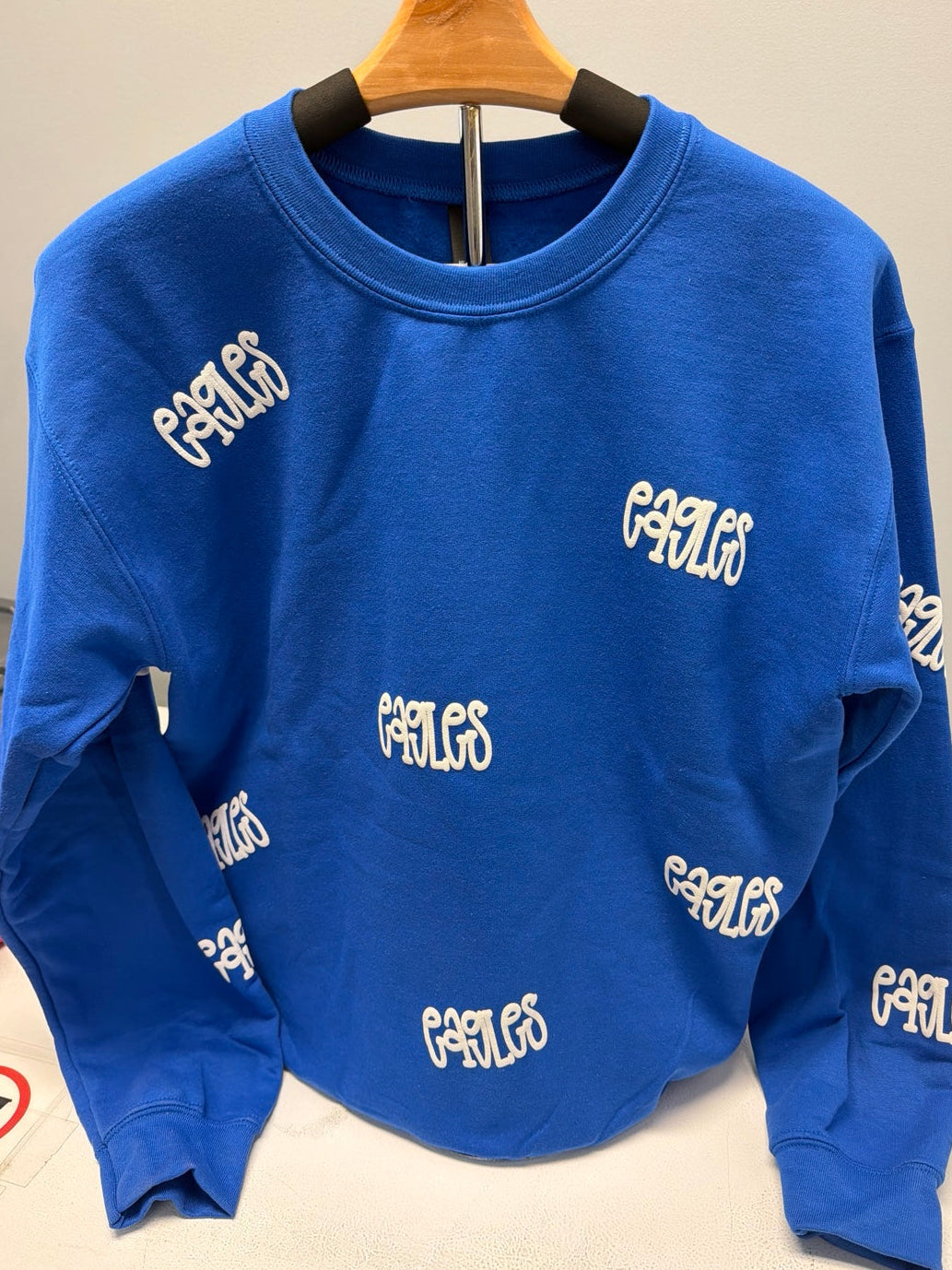 A blue crewneck sweatshirt with an all-over white 'eagles' logo print, hanging on a wooden hanger against a plain light gray wall.