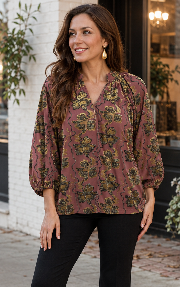 A woman stands on a city sidewalk wearing a loose, long-sleeve blouse with a muted floral pattern in dusty rose and olive green, paired with black flared pants and black ankle boots.