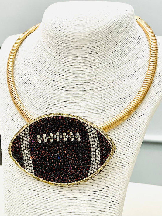 A football-shaped statement necklace with a gold-tone chain and a black rhinestone-embellished center, displayed on a white textured jewelry bust.