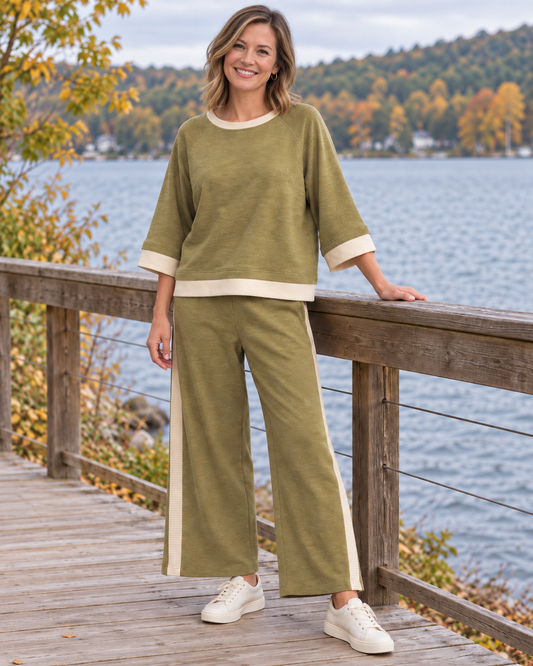 A woman stands on a wooden boardwalk by a lake wearing an olive green two-piece lounge set with cream trim, consisting of a short-sleeve top and matching wide-leg pants, paired with white sneakers.