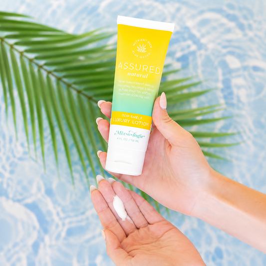Hand holding a tube of Assured lotion with a palm leaf in the background