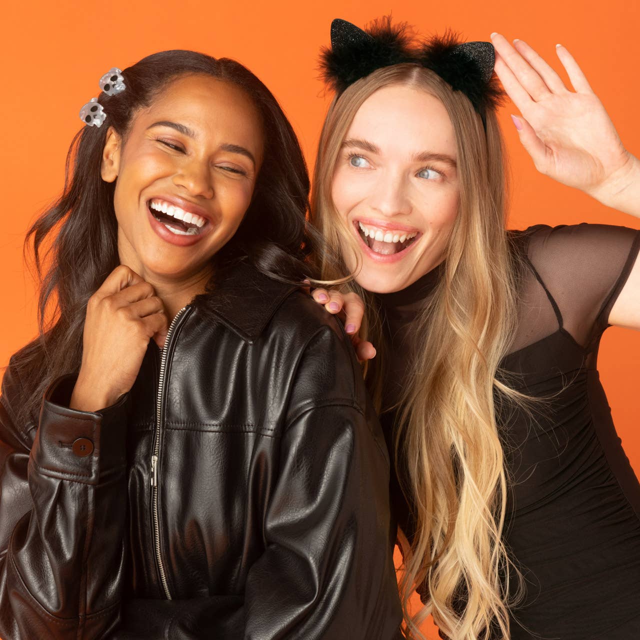 Two women laughing closely against a solid orange background. The woman on the left wears a black faux-leather jacket and a skull hair clip, while the woman on the right wears black cat ears and a black sheer-sleeve top, waving her hand.