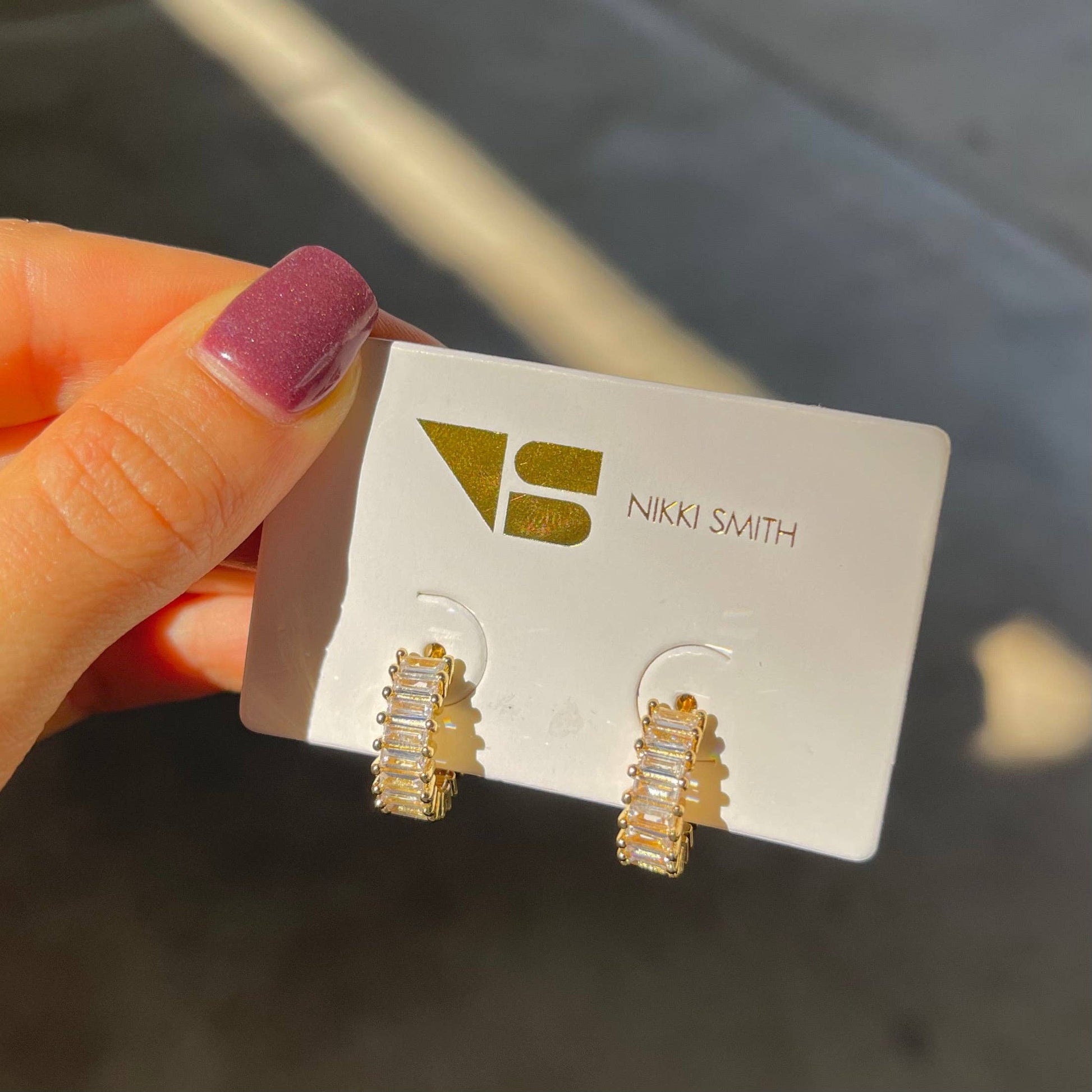 Gold earrings on a Nikki Smith card held by a hand with purple nail polish.