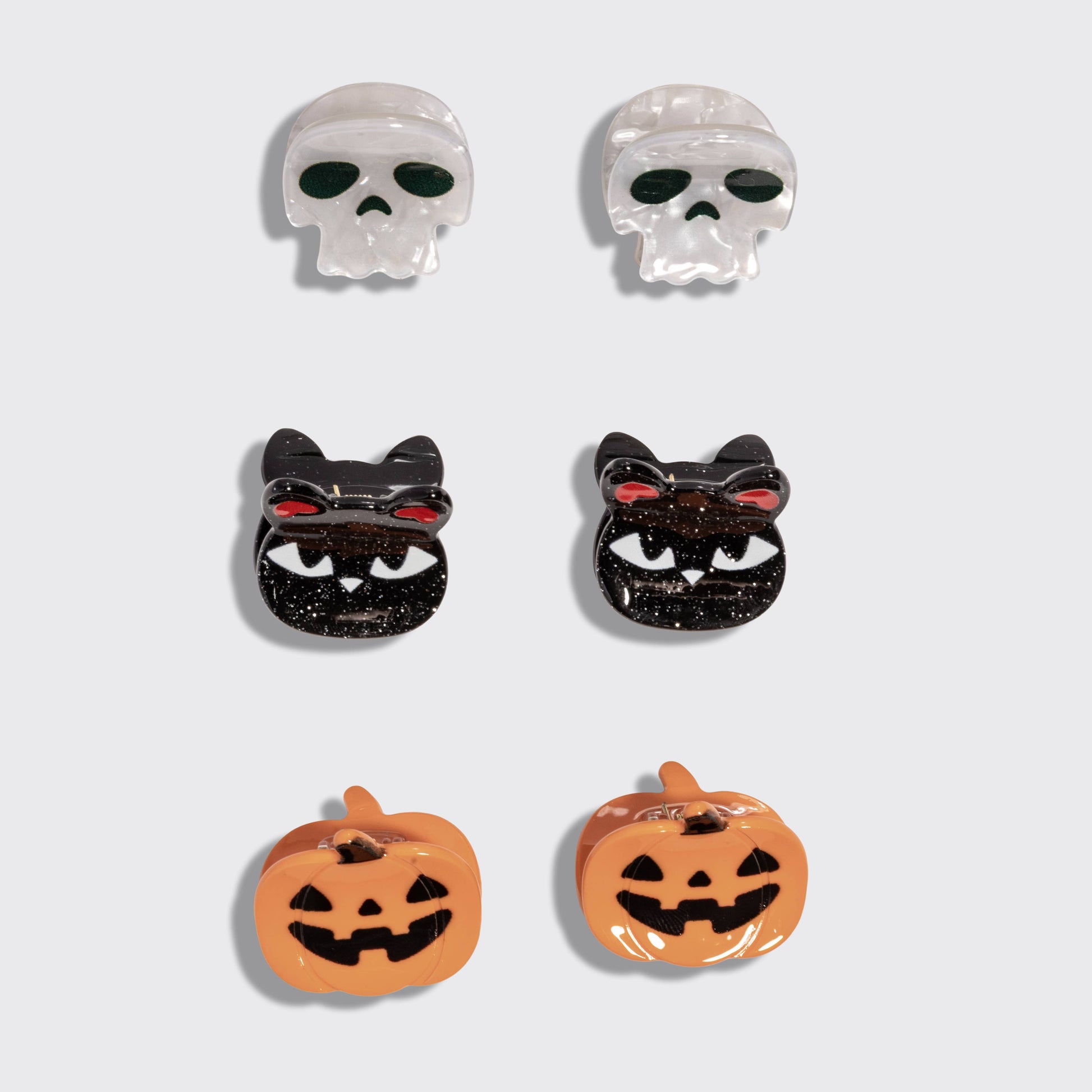 Six Halloween-themed stud earrings arranged in a 3-by-2 grid on a light gray background. The set includes two white skulls with dark green eyes, two black cats with red eyes and white teeth, two orange pumpkins with black jack-o'-lantern faces, and two clear skull earrings with dark green eyes.