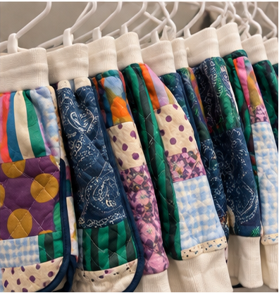 A close-up of several quilted patchwork sweatshirt sleeves hanging on white hangers. The sleeves feature a variety of colorful patterns including paisley, gingham, polka dots, stripes, and floral prints in shades of blue, green, purple, pink, orange, and cream.