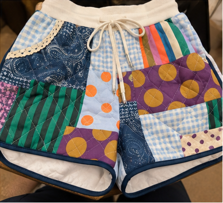 A pair of handmade patchwork quilted shorts featuring a variety of colorful fabric panels including gingham, paisley, stripes, polka dots, and solid colors, with a cream drawstring waist.