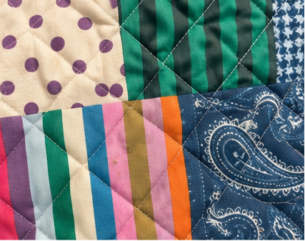 Close-up of a quilted patchwork fabric surface with multiple textile patterns: purple polka dots on cream, green and dark green vertical stripes, blue and white vertical stripes, pink and tan stripes, and a blue paisley print.
