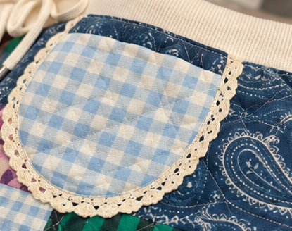 Close-up of a handmade quilted apron or pouch featuring a light blue and white gingham fabric pocket with a cream lace trim, layered over a blue paisley quilted fabric.