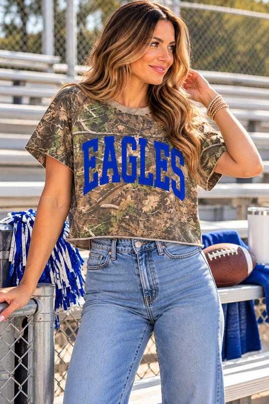 A woman stands in a gym or stadium setting wearing a camouflage graphic t-shirt with 'EAGLES' printed in bold blue letters across the chest, paired with light blue jeans.