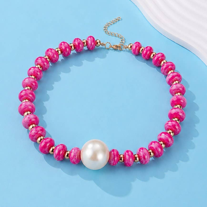 A beaded necklace featuring textured hot pink marbled beads, small gold spacer beads, a large white pearl at the center, and a gold-tone chain clasp, displayed on a light blue background.