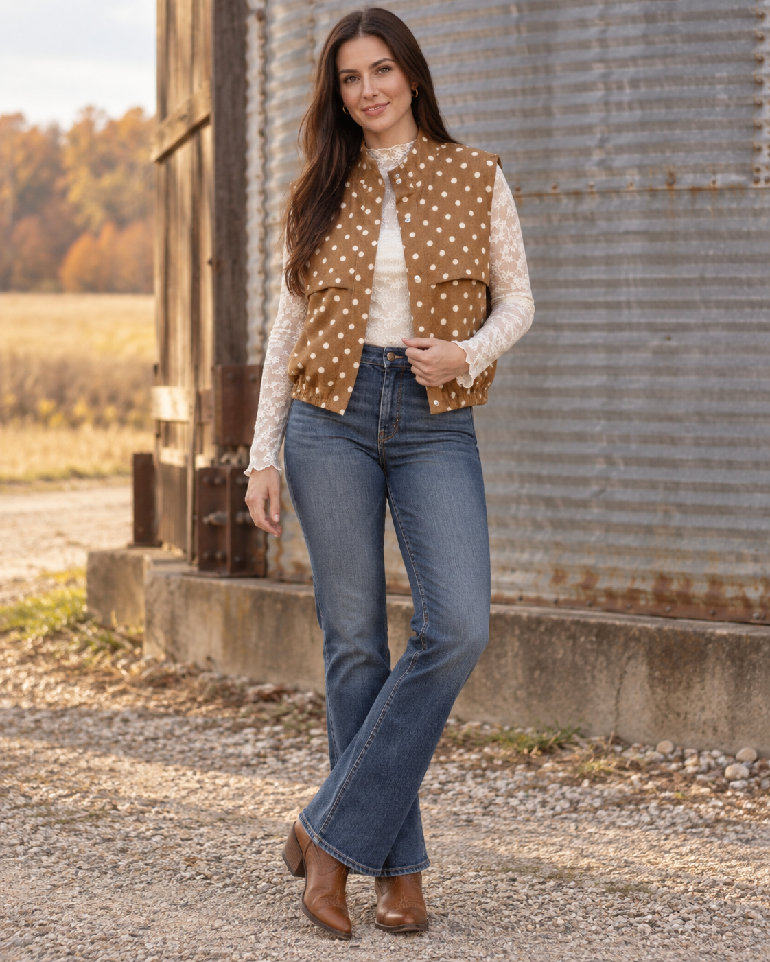 A woman stands outdoors in front of a rustic corrugated metal building, wearing a brown vest with white polka dots over a cream long-sleeve top, blue jeans, and brown ankle boots.