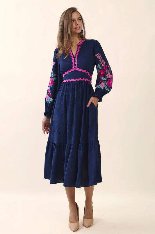 Woman wearing a navy embroidered tiered midi dress with floral long sleeves and pink ric rac trim.