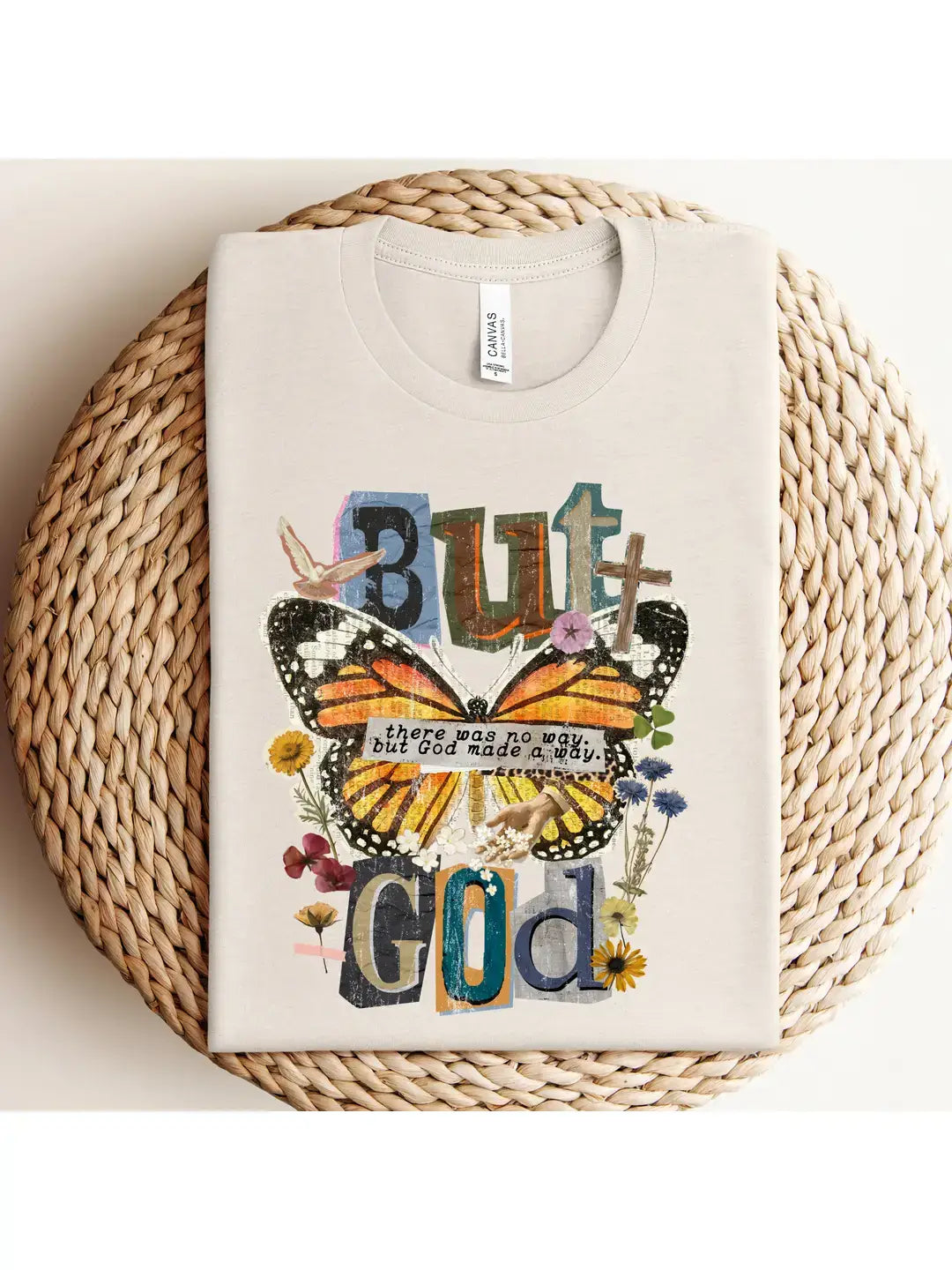 A folded cream-colored t-shirt with a colorful graphic print featuring a butterfly, flowers, and the text 'But God' on a round woven straw mat.
