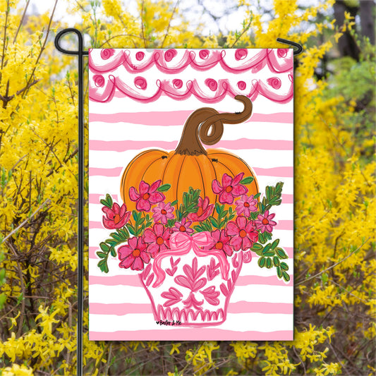 Decorative garden flag with a pumpkin and floral design on a pink and white striped background, set against a yellow flowering bush.