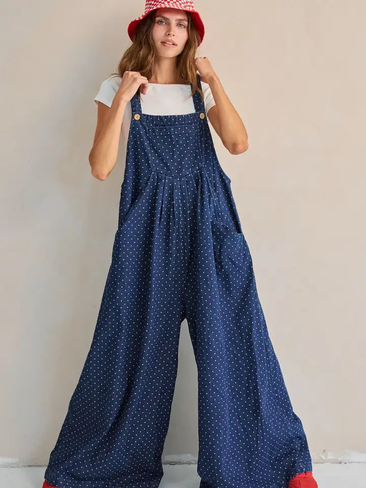 Navy polka dot wide-leg overall jumpsuit, standing front view with hand on hat brim