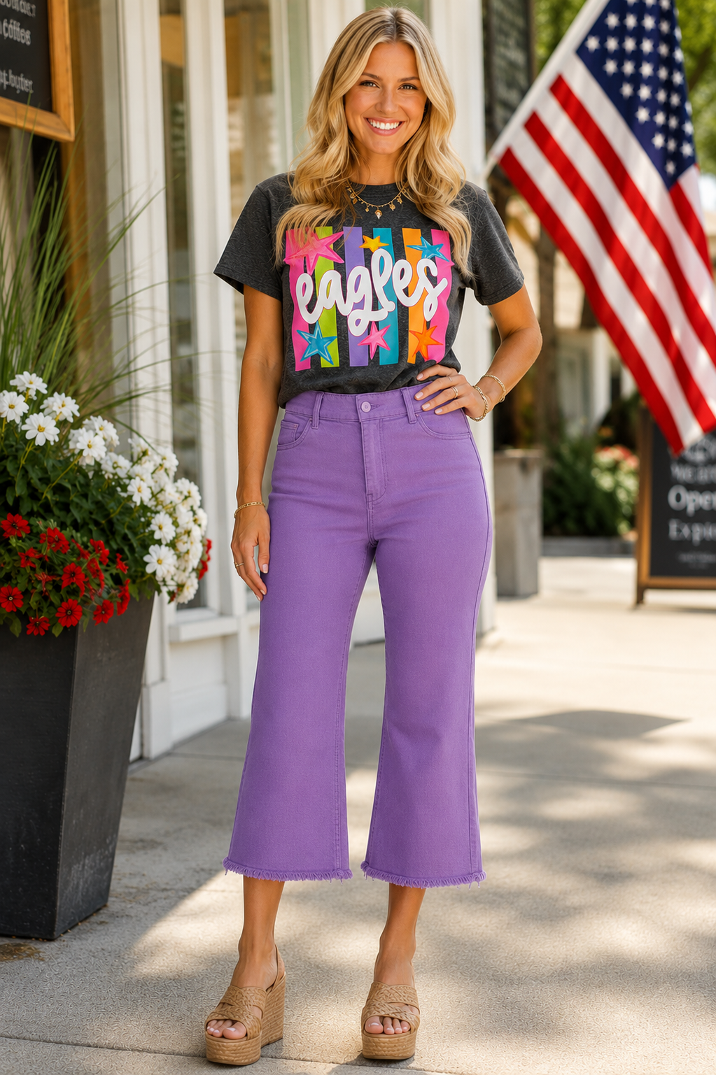 A woman modeling a black graphic t-shirt with a colorful 'Eagles' design and purple cropped flare jeans, standing outdoors on a sidewalk in front of a storefront with an American flag in the background.