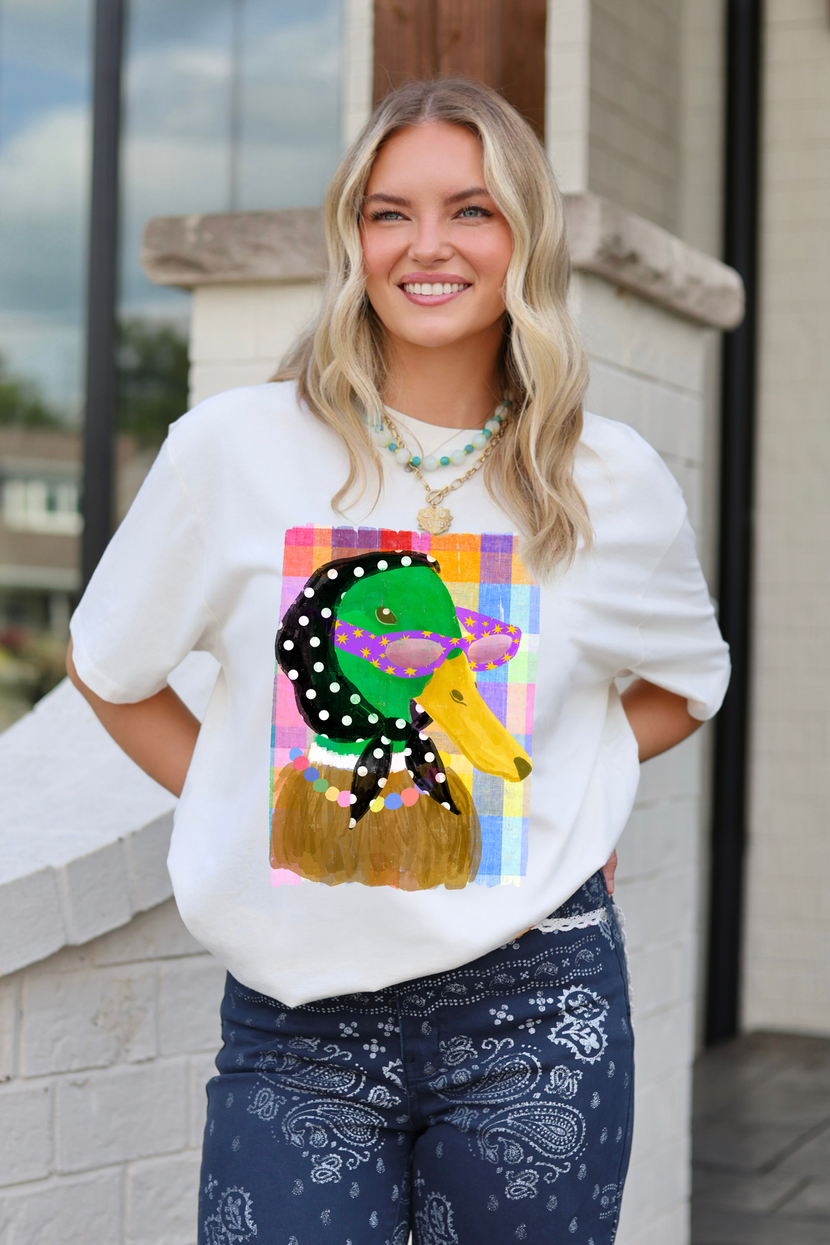 A smiling blonde woman wearing a white graphic poppy & pine t-shirt with a colorful cartoon duck, paired with blue paisley bandana jeans, standing outdoors in front of a light-colored brick wall.