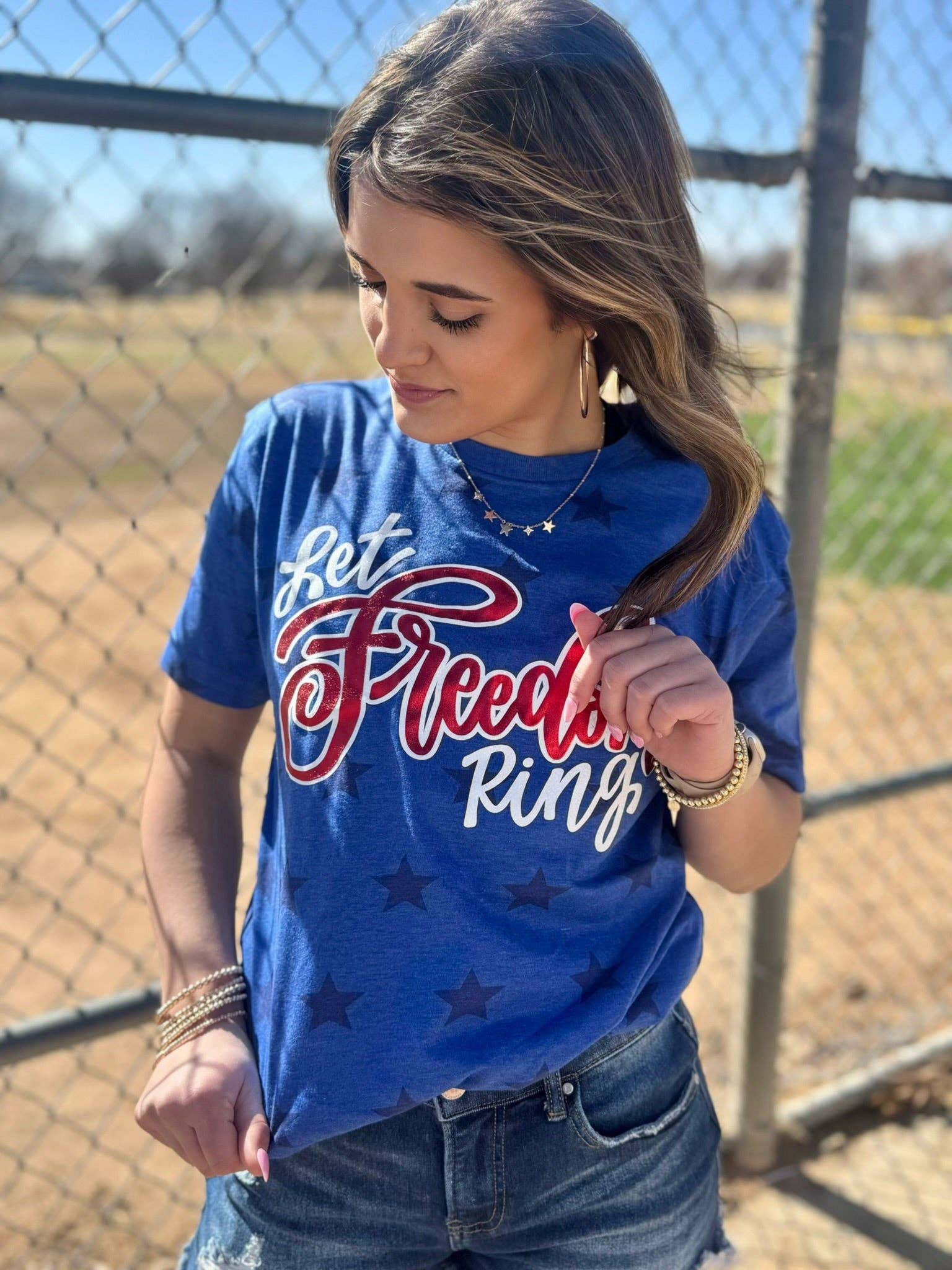 Woman wearing a blue t-shirt with 'Let Freedom Ring' text, standing in front of a chain-link fence.