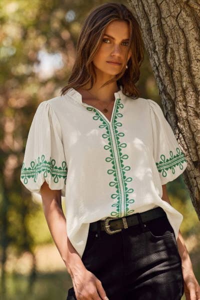 Woman wearing a white blouse with green embroidery standing against a tree in a natural setting