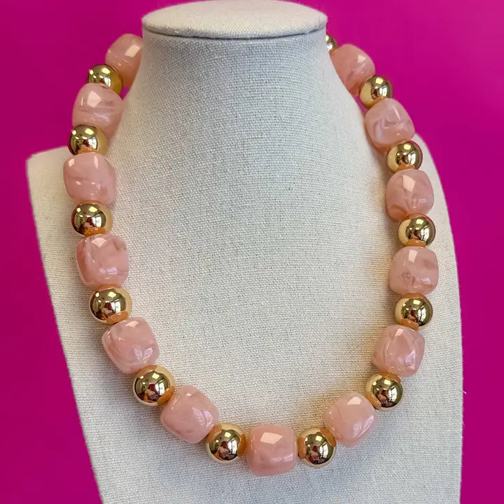 A pink beaded necklace with faceted pink stone beads and shiny gold metallic spacer beads, displayed on a white fabric-covered jewelry bust against a bright pink background.