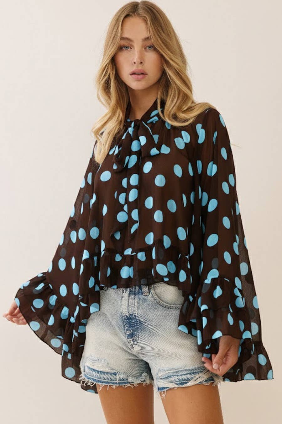 Woman wearing a brown blouse with blue polka dots and denim shorts on a plain background