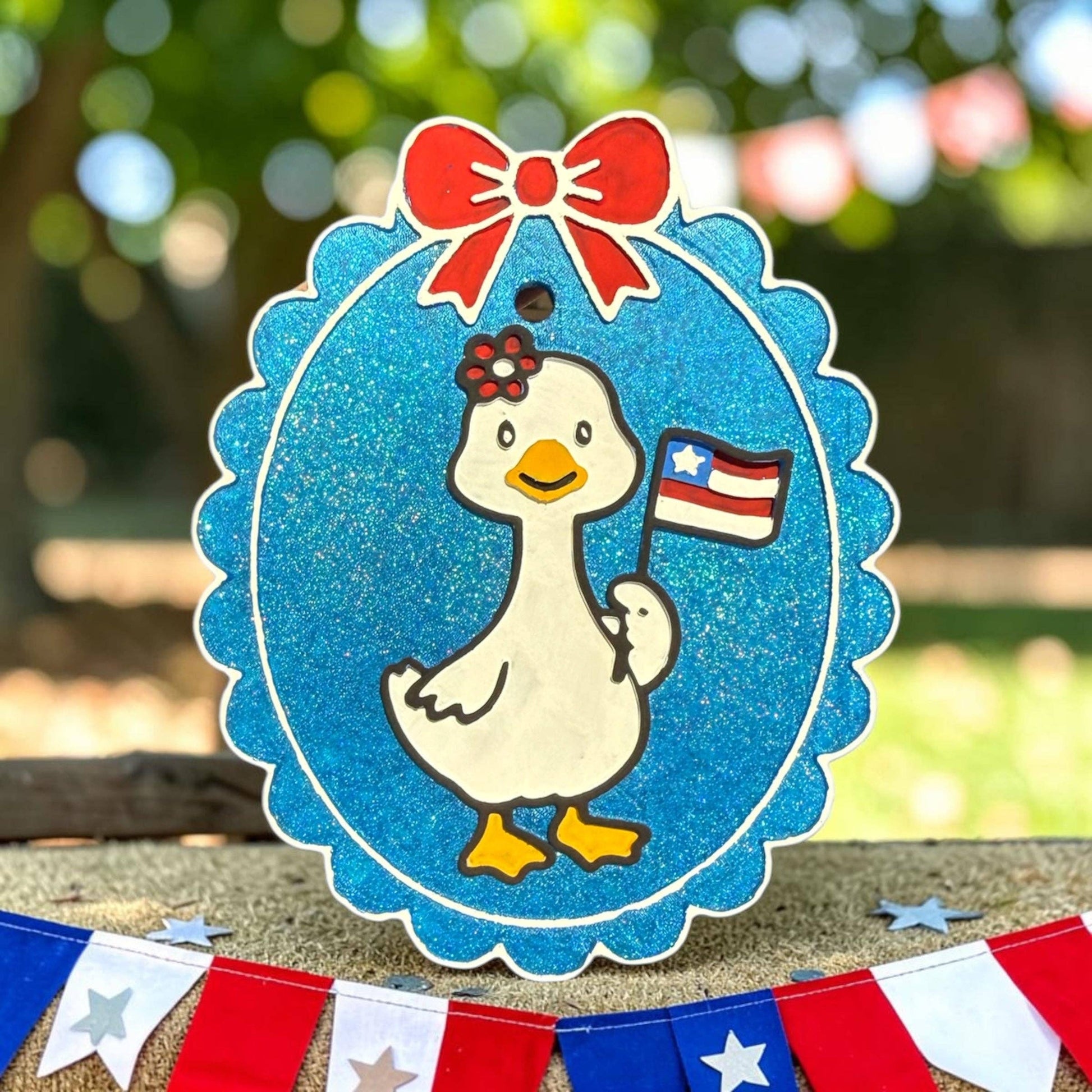 Decorative sign with a goose holding an American flag, surrounded by a blue border with a red bow, on a blurred outdoor background.