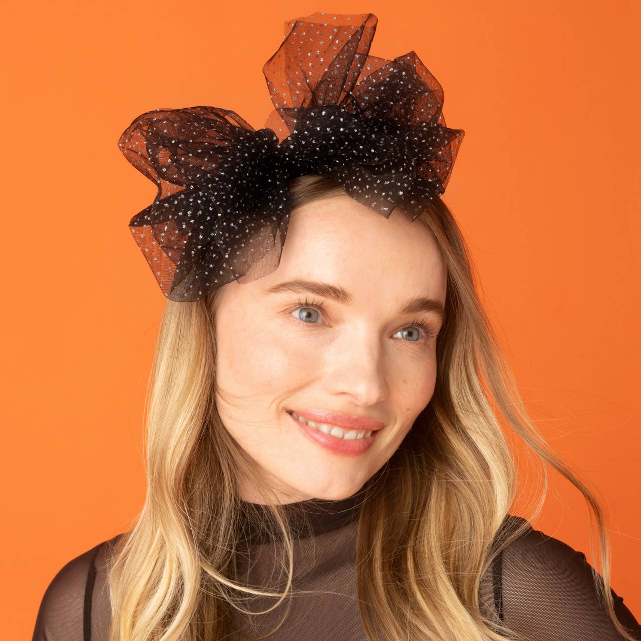 A woman with blonde hair wearing a large black sheer polka-dot fascinator headband, set against a solid orange background.