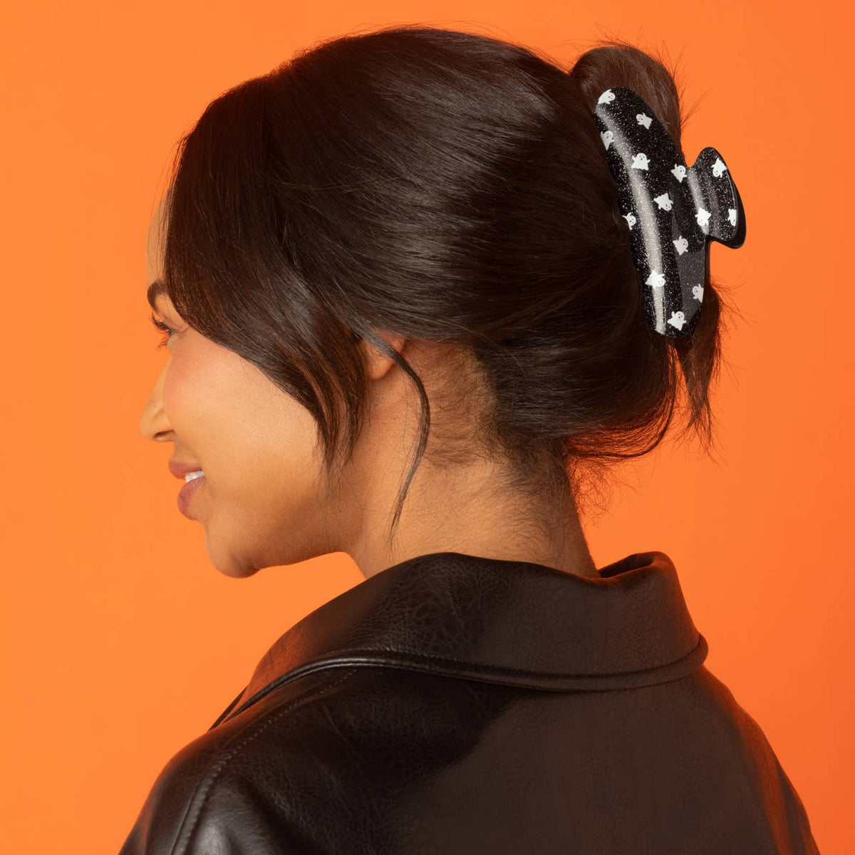 A woman in profile wearing a black hair claw clip decorated with white stars, set against a solid orange background.