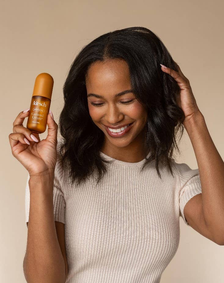 A woman with dark wavy hair smiles while holding a small amber bottle labeled koch cosmetics in her right hand, against a plain beige studio background.