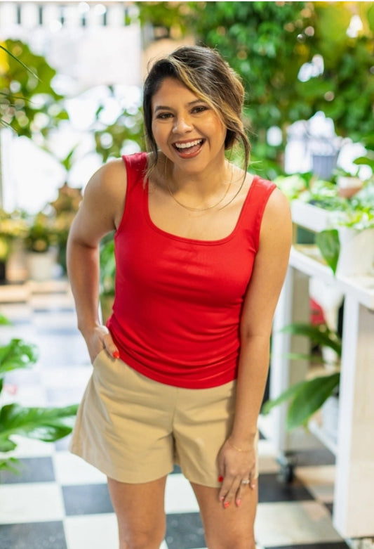 Woman wearing a red tank top and beige shorts standing outdoors with greenery in the background