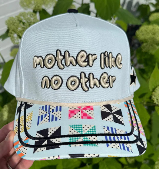 A light blue baseball cap with 'mother like no other' embroidered on the front, held by a hand against a background of green leaves and flowers. The brim features a colorful geometric pinwheel pattern.